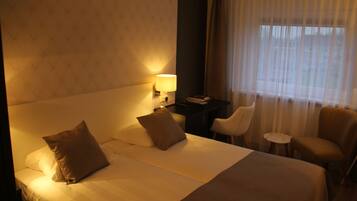 Habitación de hotel con una cama grande, dos almohadas, un escritorio con lámpara, una silla y vistas al exterior a través de cortinas traslúcidas.