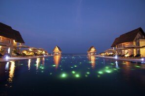 Una piscina con estructuras de techo de paja a ambos lados, reflejos de luces en el agua.