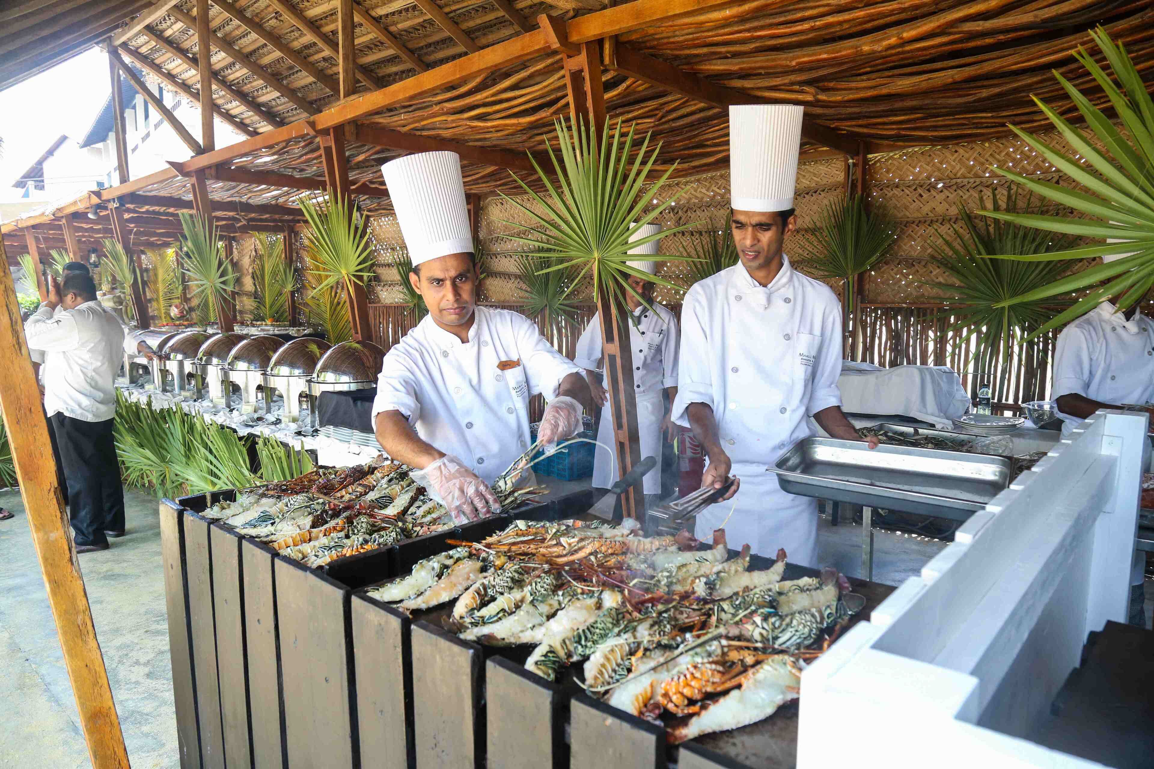 Chefs preparing seafood at an outdoor buffet with a wooden structure and palm trees in the background.