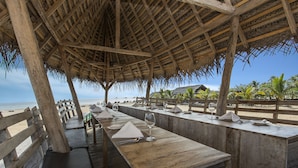 An outdoor dining area with wooden tables and benches, a thatched roof, and a view of the beach and ocean. - Maalu Maalu Resort & Spa (Pasikuda)