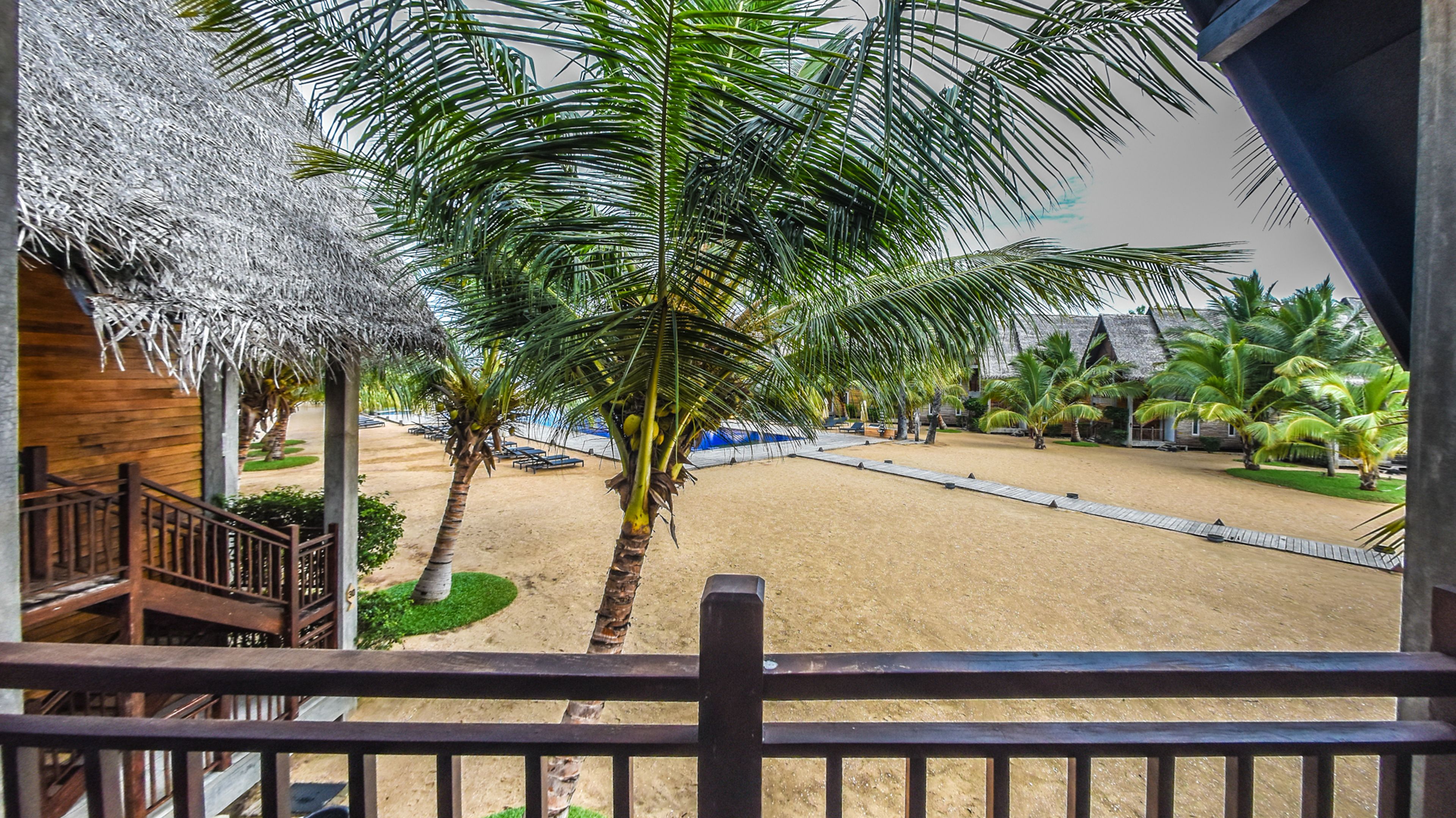 A beach view from a balcony with palm trees, a sandy beach, and a thatched-roof building.