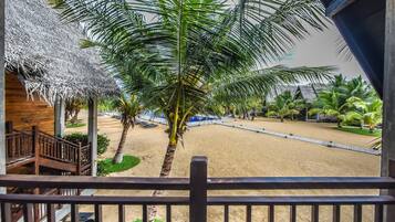 A beach view from a balcony with palm trees, a sandy beach, and a thatched-roof building.