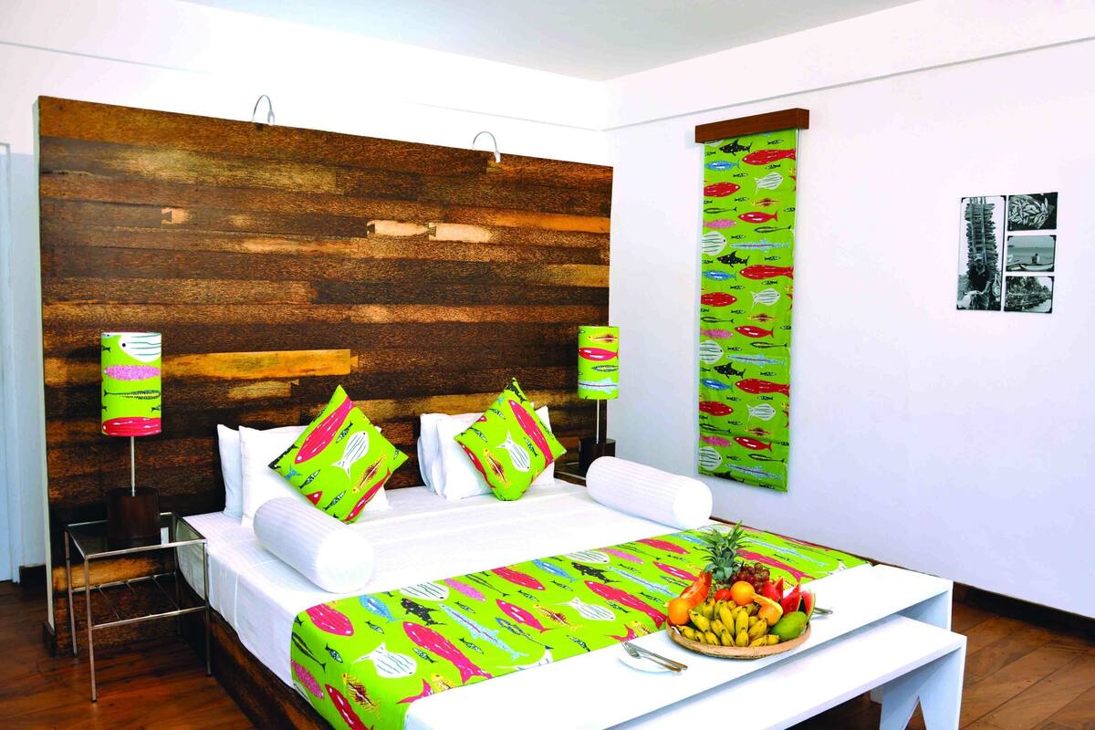 Habitación de hotel con cabecera de madera, cama con sábanas blancas, almohadas coloridas y un frutero en la mesita de noche.