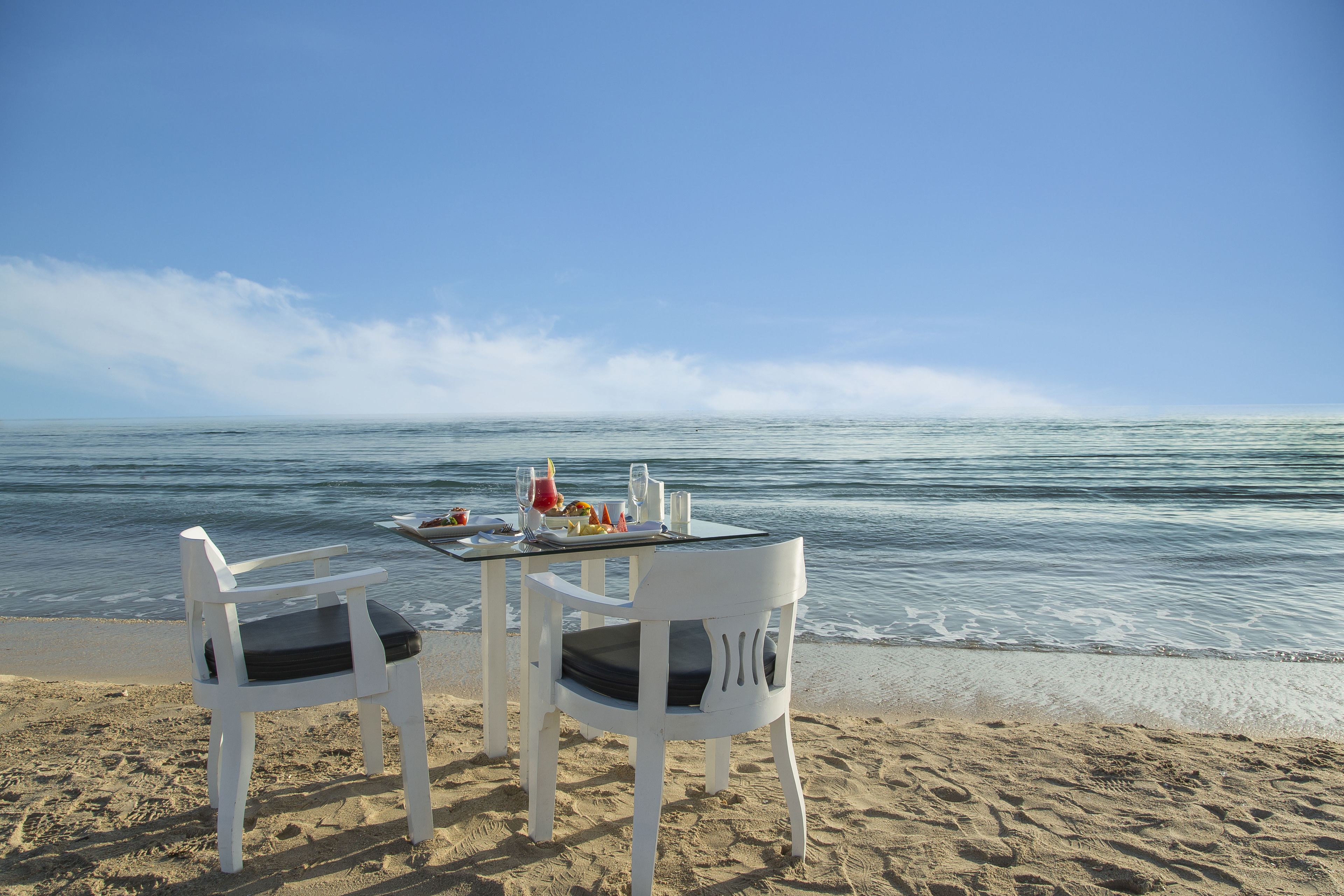 A beach table with two white chairs, a glass top, and a meal set.