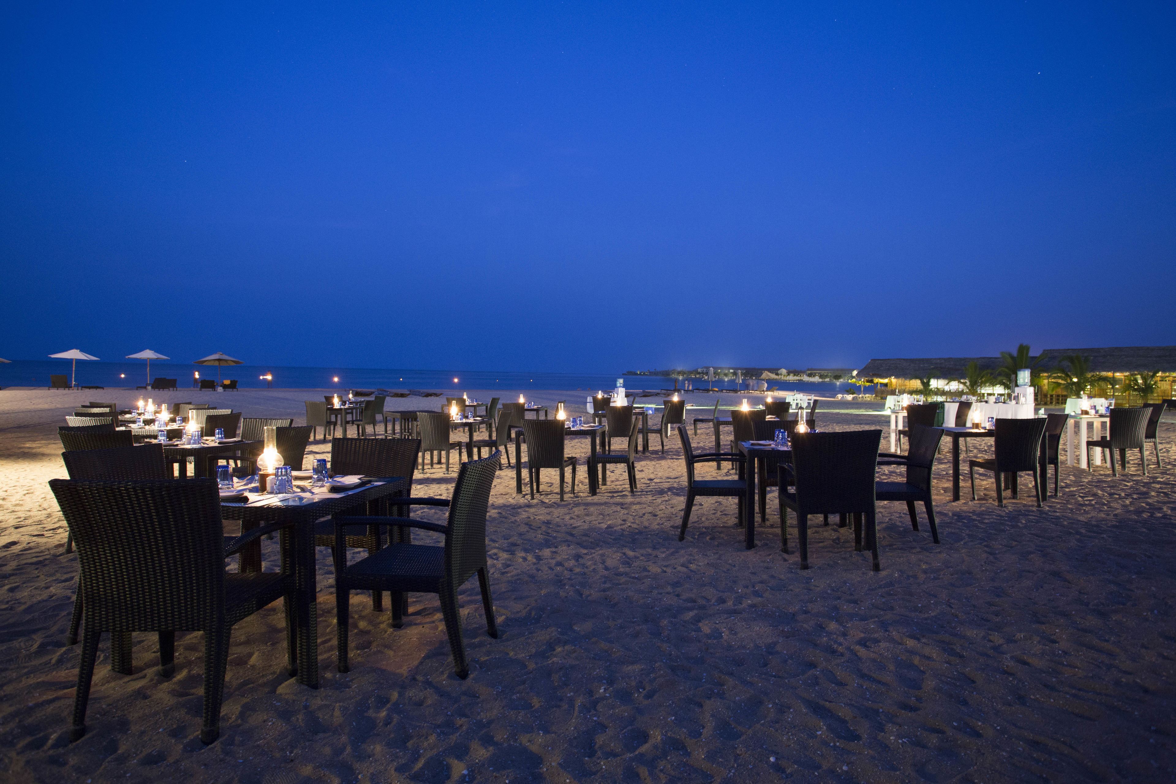Un espace de restauration en plein air, avec des tables et des chaises installées sur une plage de sable, offrant une vue sur l’océan.