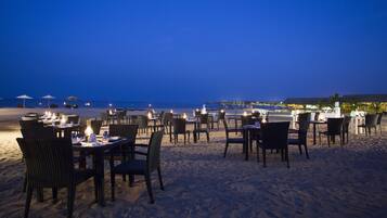 Un espace de restauration en plein air, avec des tables et des chaises installées sur une plage de sable, offrant une vue sur l’océan.