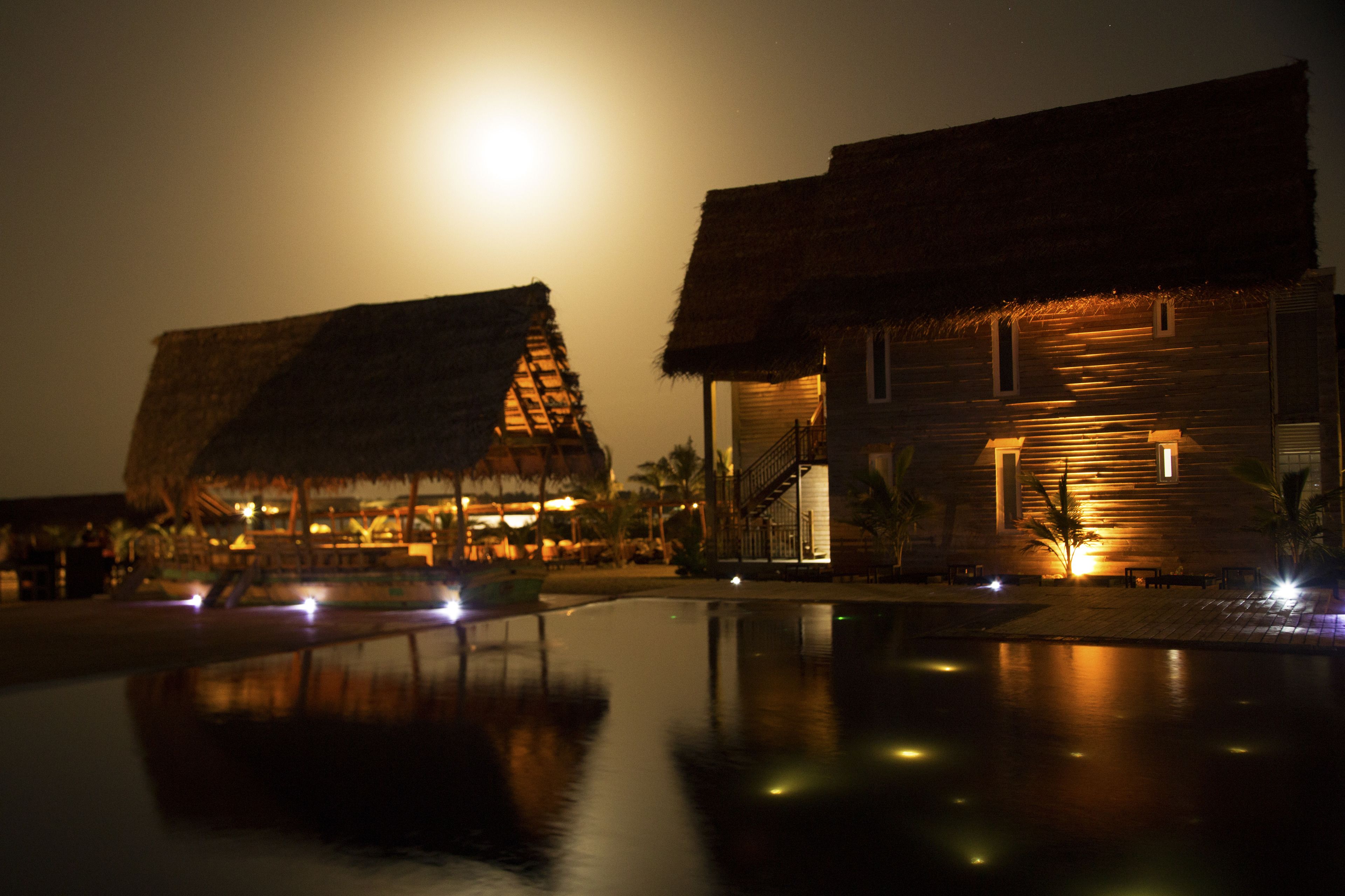 Um resort com construções de telhado de palha, piscina e céu iluminado pela lua.