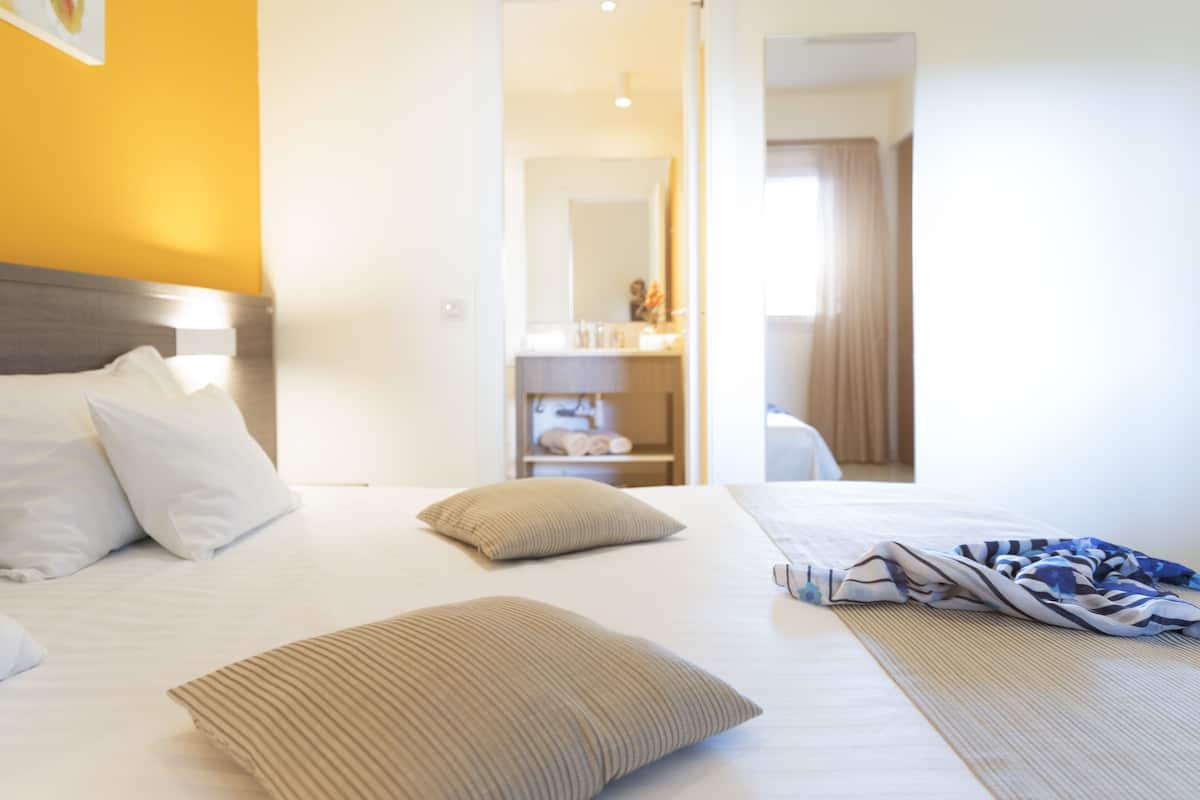 Una habitación de hotel con una cama, almohadas, una mesita de noche y un baño visible a través de una puerta abierta.