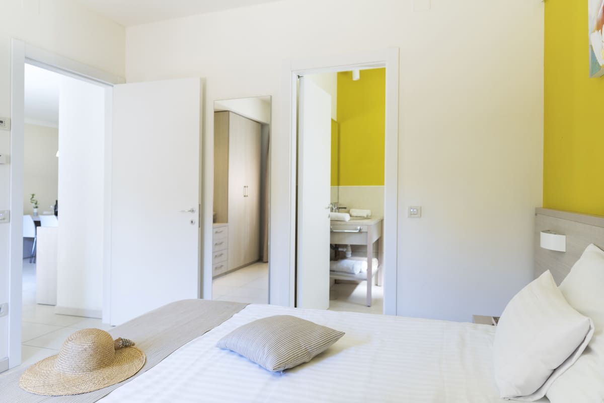 Una habitación de hotel con una cama, un sombrero sobre la cama, un baño con un espejo y una pared amarilla.