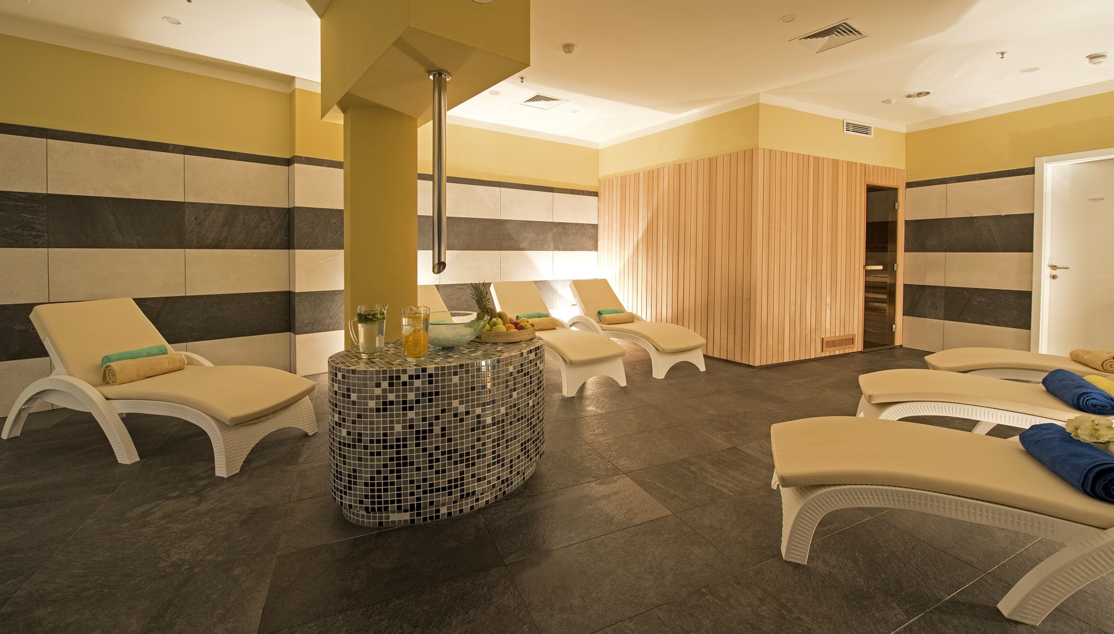 Um espaço de spa com várias poltronas confortáveis, piso revestido e uma mesa central com uma cesta de frutas e um jarro.
