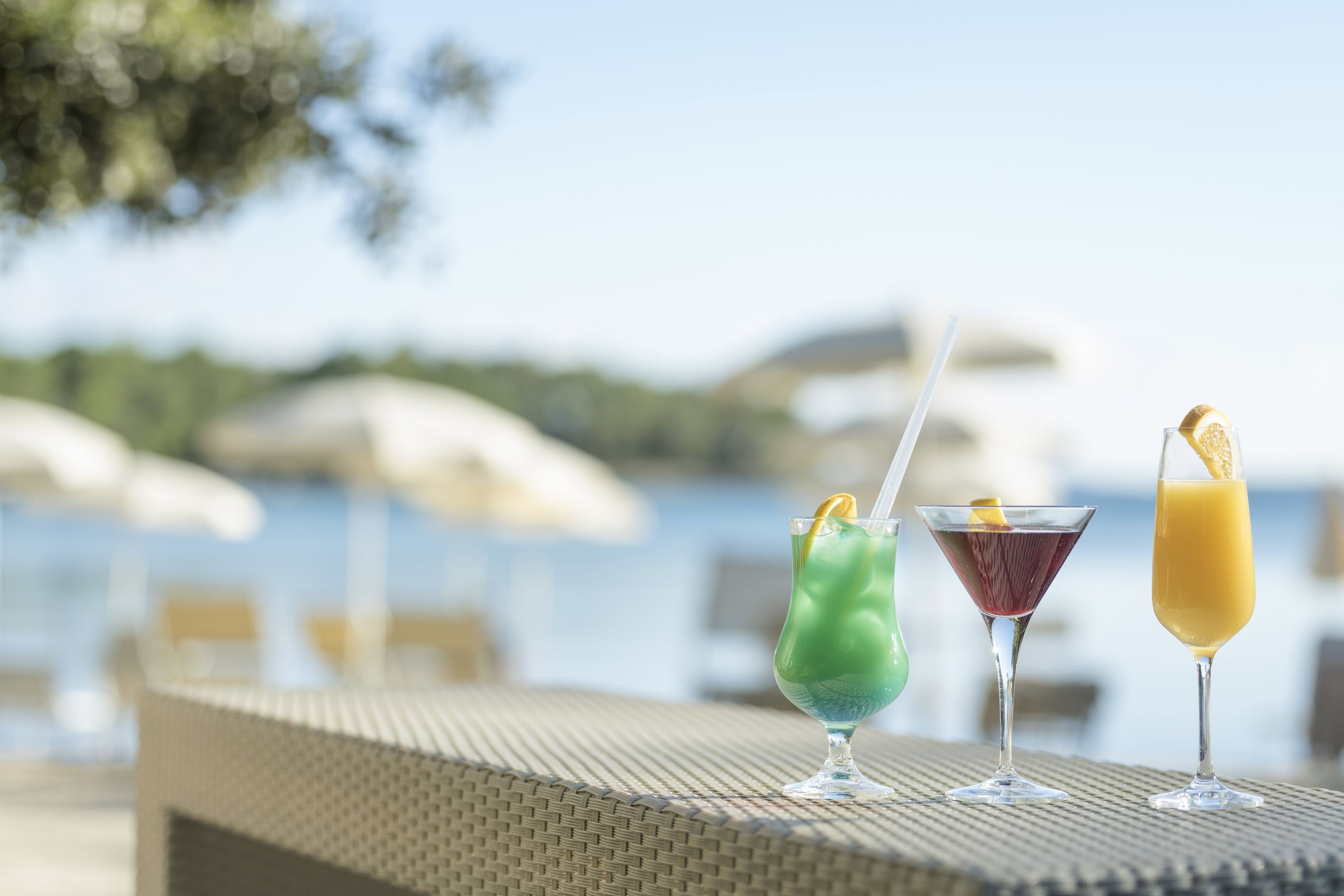 Three cocktails on a table with a view of the sea and umbrellas in the background.
