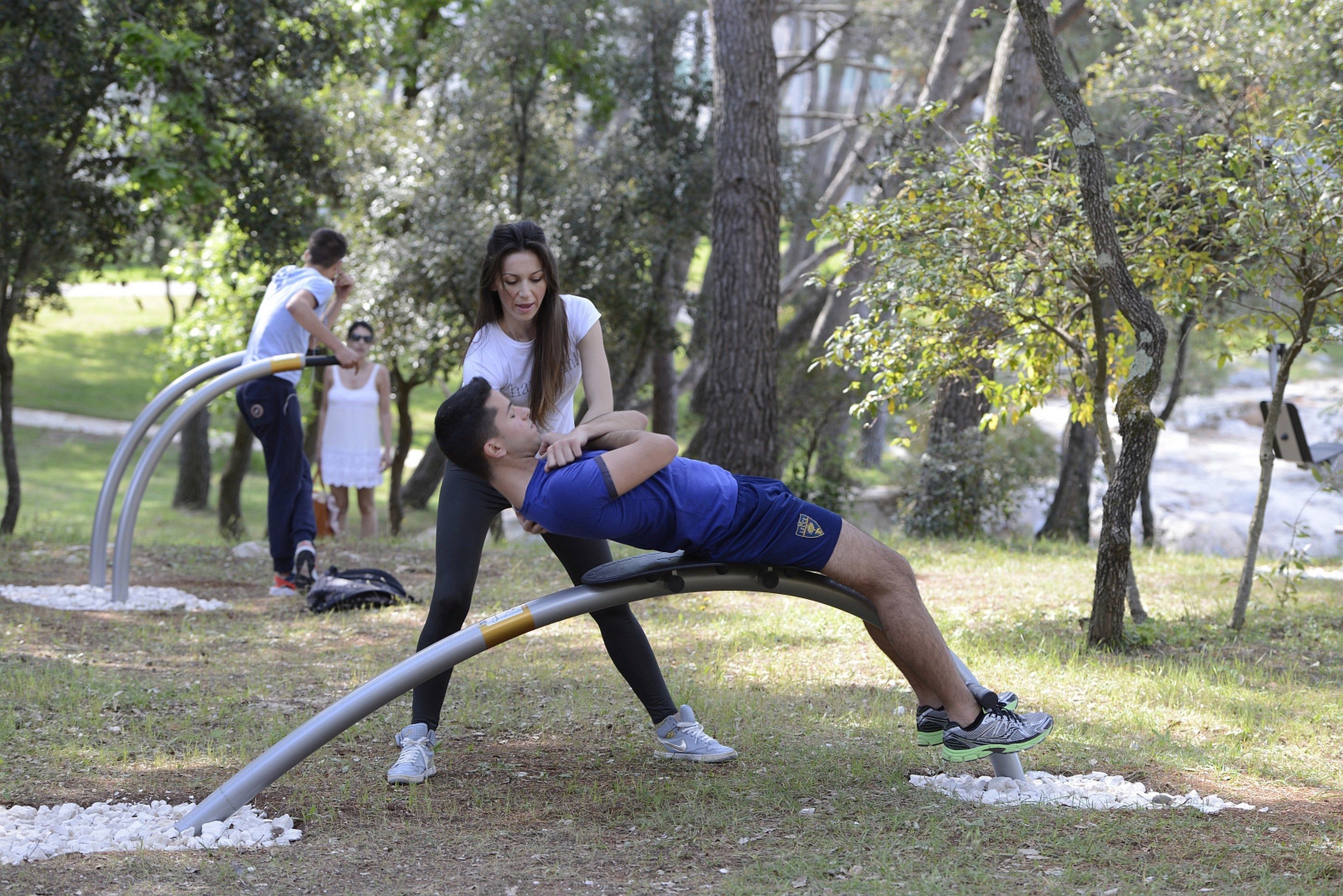 A person performing a stretching exercise on a curved metal bar in an outdoor setting with trees and grass.