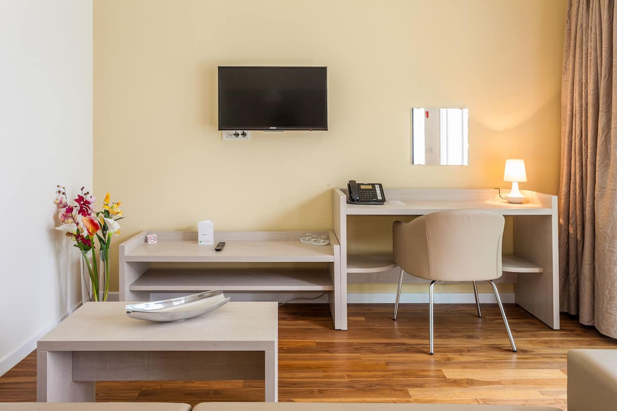 Habitación de hotel con televisor de pantalla plana en la pared, escritorio con teléfono, una silla, una mesita con un cuenco decorativo y un florero con flores.