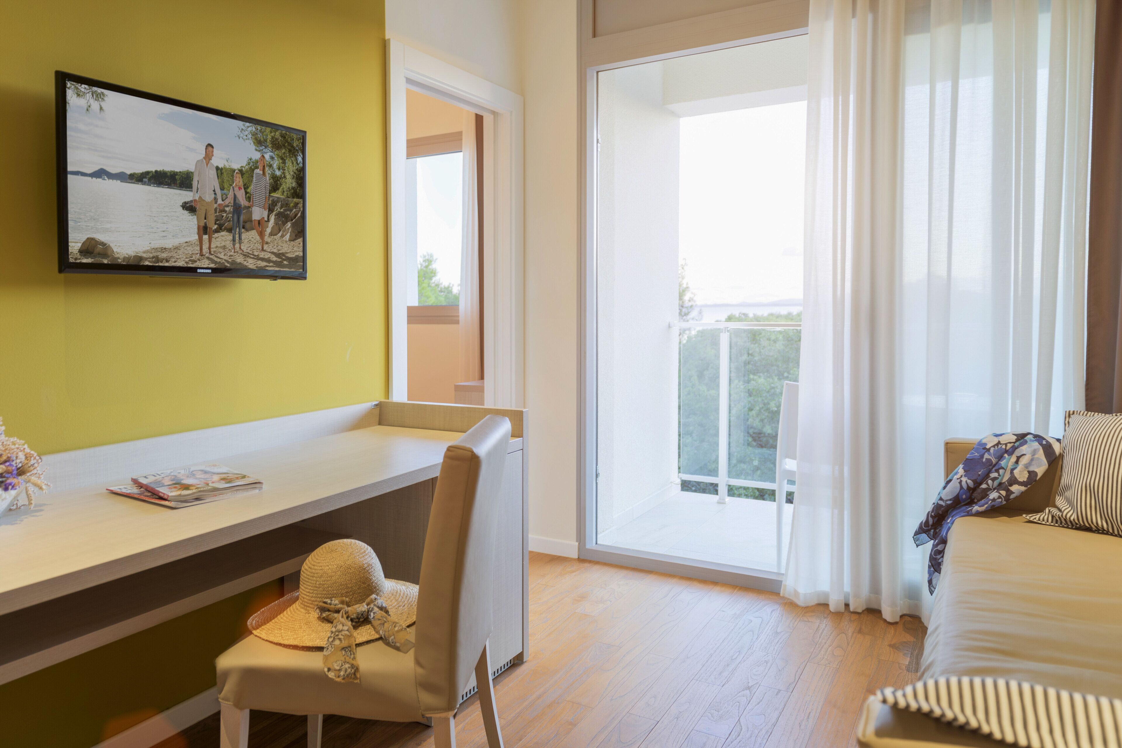 A bedroom with a bed, a desk with a chair, a television, and a large window with curtains.