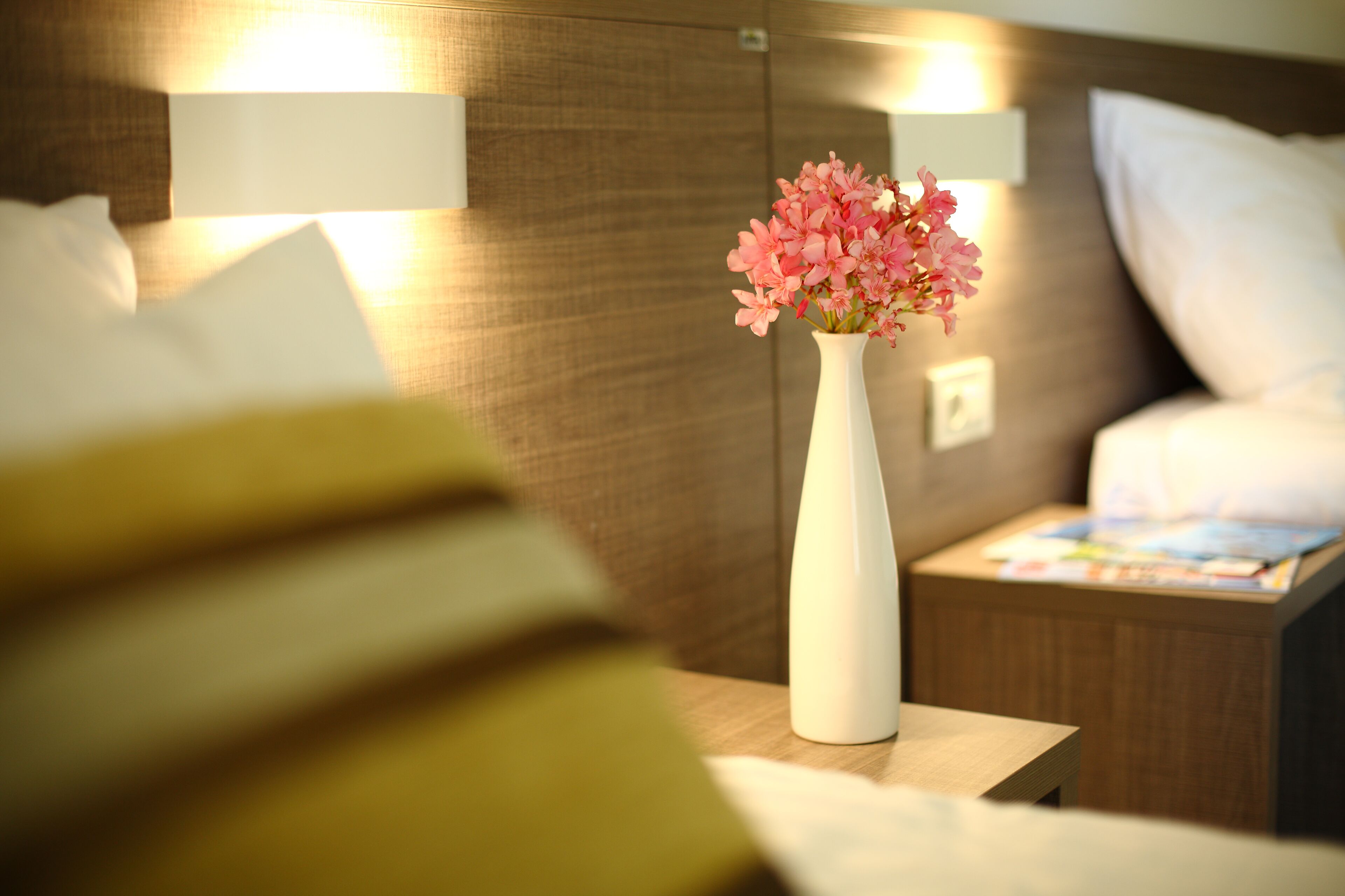 A hotel room with a white vase of pink flowers on a bedside table, a wooden headboard, and a nightstand with a lamp.