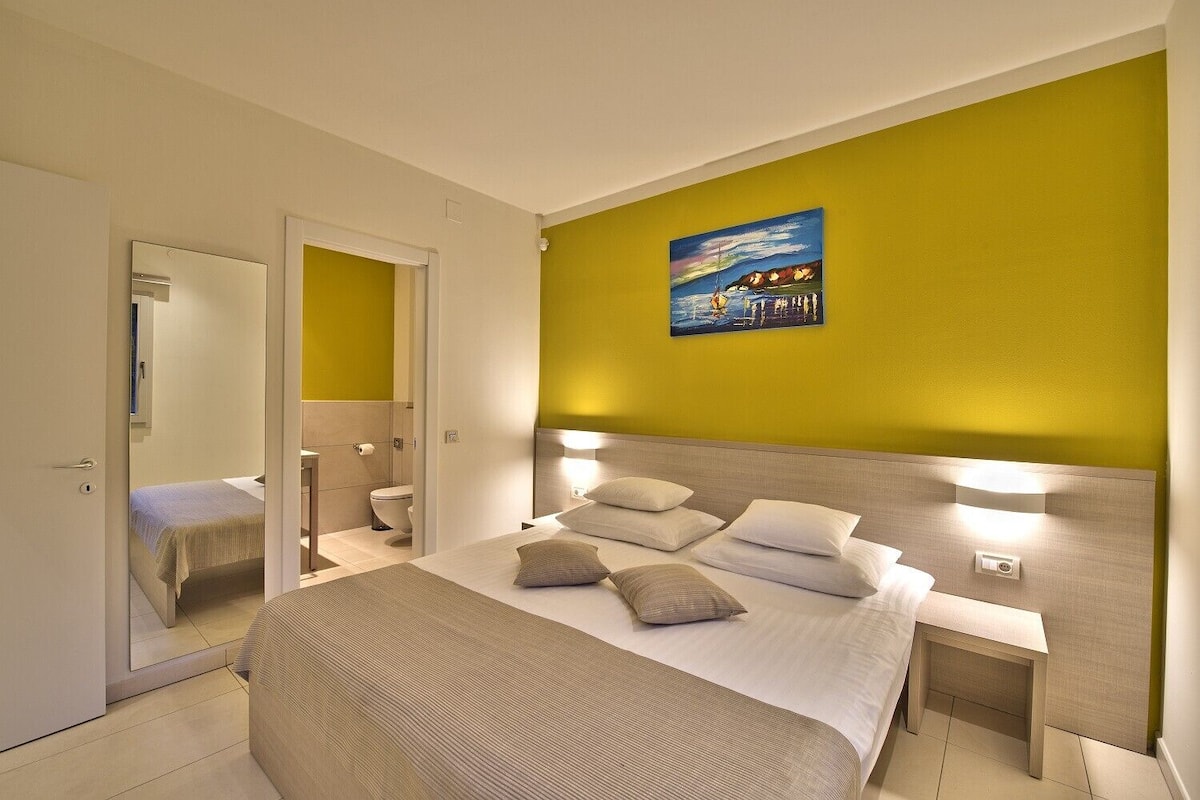 Habitación de hotel con cama, mesitas de noche, un espejo, un cuadro en la pared y un baño visible a través de una puerta abierta.