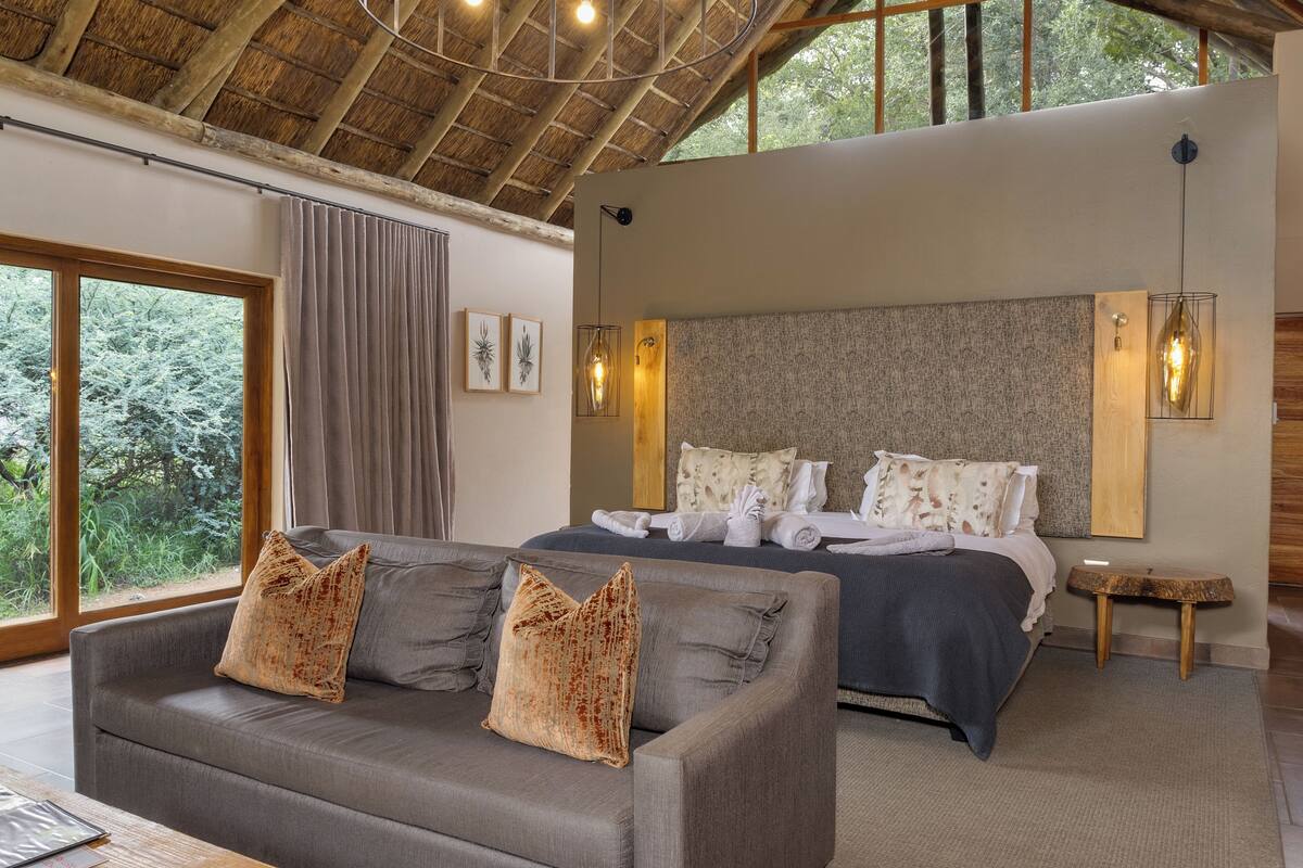 A modern bedroom with a large bed, a sofa, and a view of trees through the window.