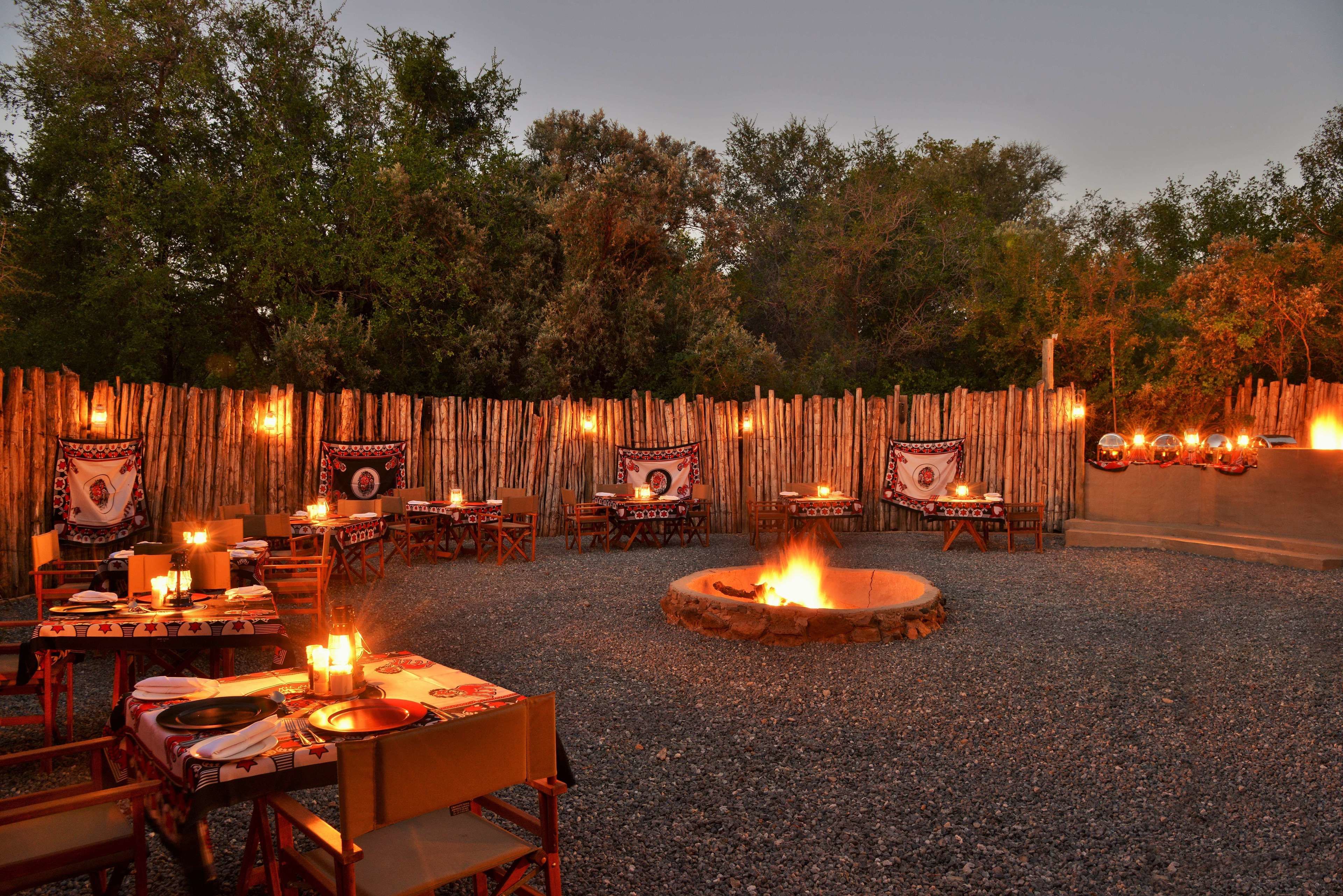 An outdoor dining area with tables set for a meal, surrounded by a wooden fence, and a central fire pit.