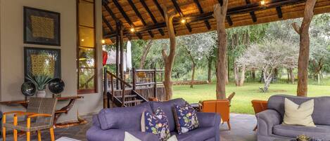 A covered outdoor seating area with a sofa and chairs, a staircase, and a view of the surrounding trees.