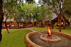 An outdoor area with a fire pit surrounded by stone, a grassy lawn, and a building with multiple rooms and tables. - Black Rhino Game Lodge (Pilanesberg National Park)