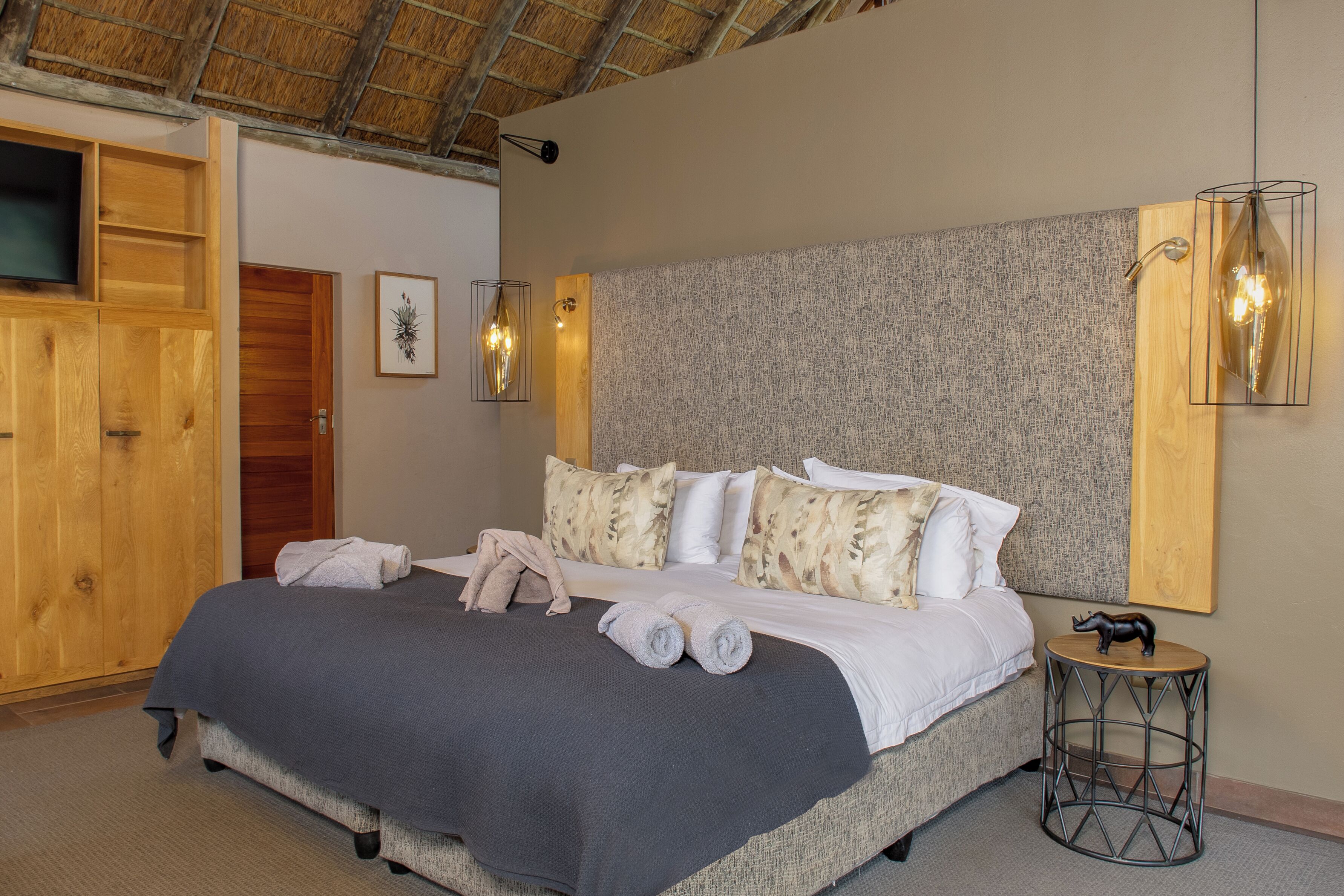 A neatly made bed with a grey blanket and rolled towels, a wooden wardrobe, a framed artwork, and two wall-mounted lamps.