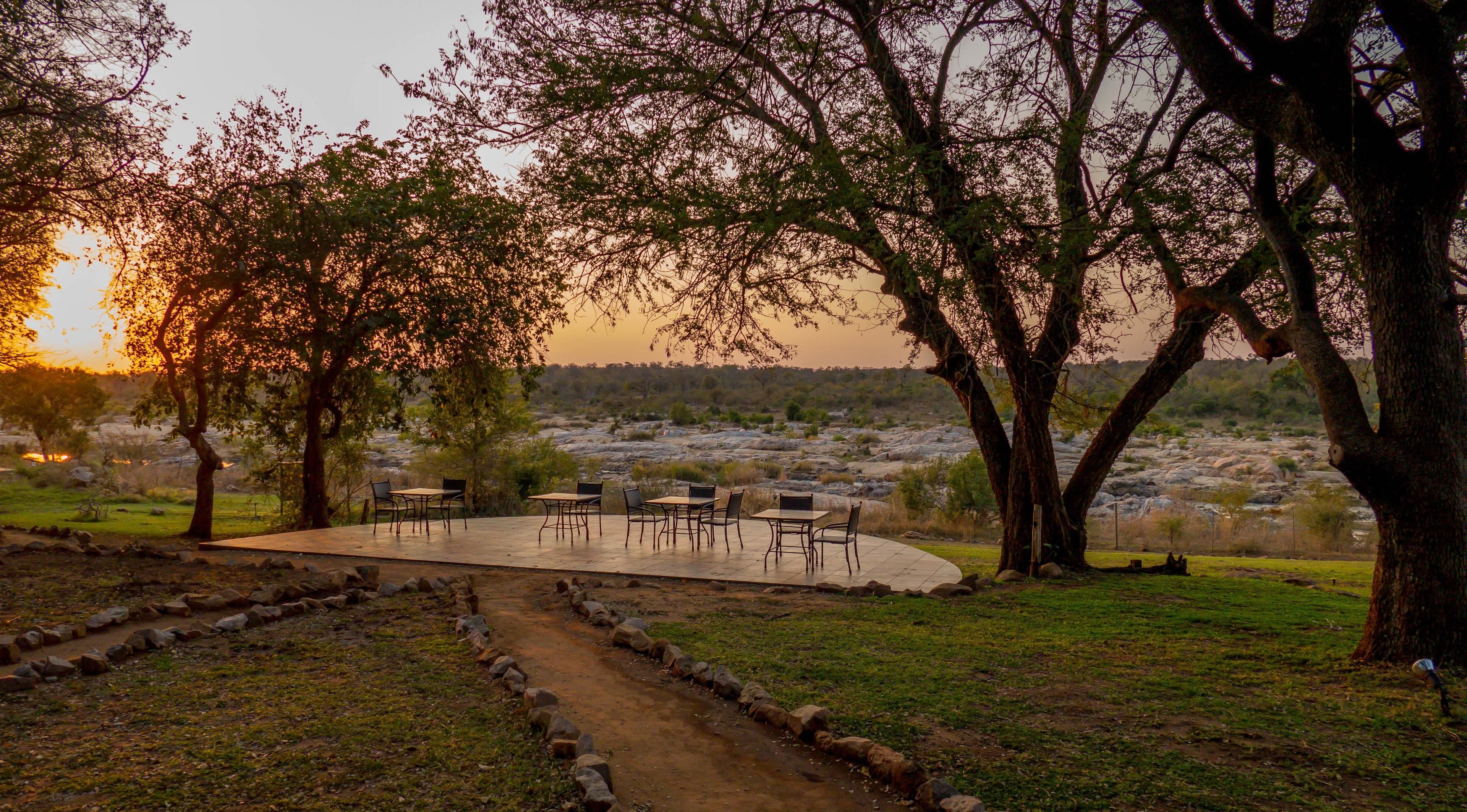 An outdoor seating area with tables and chairs, surrounded by trees and a view of a river.