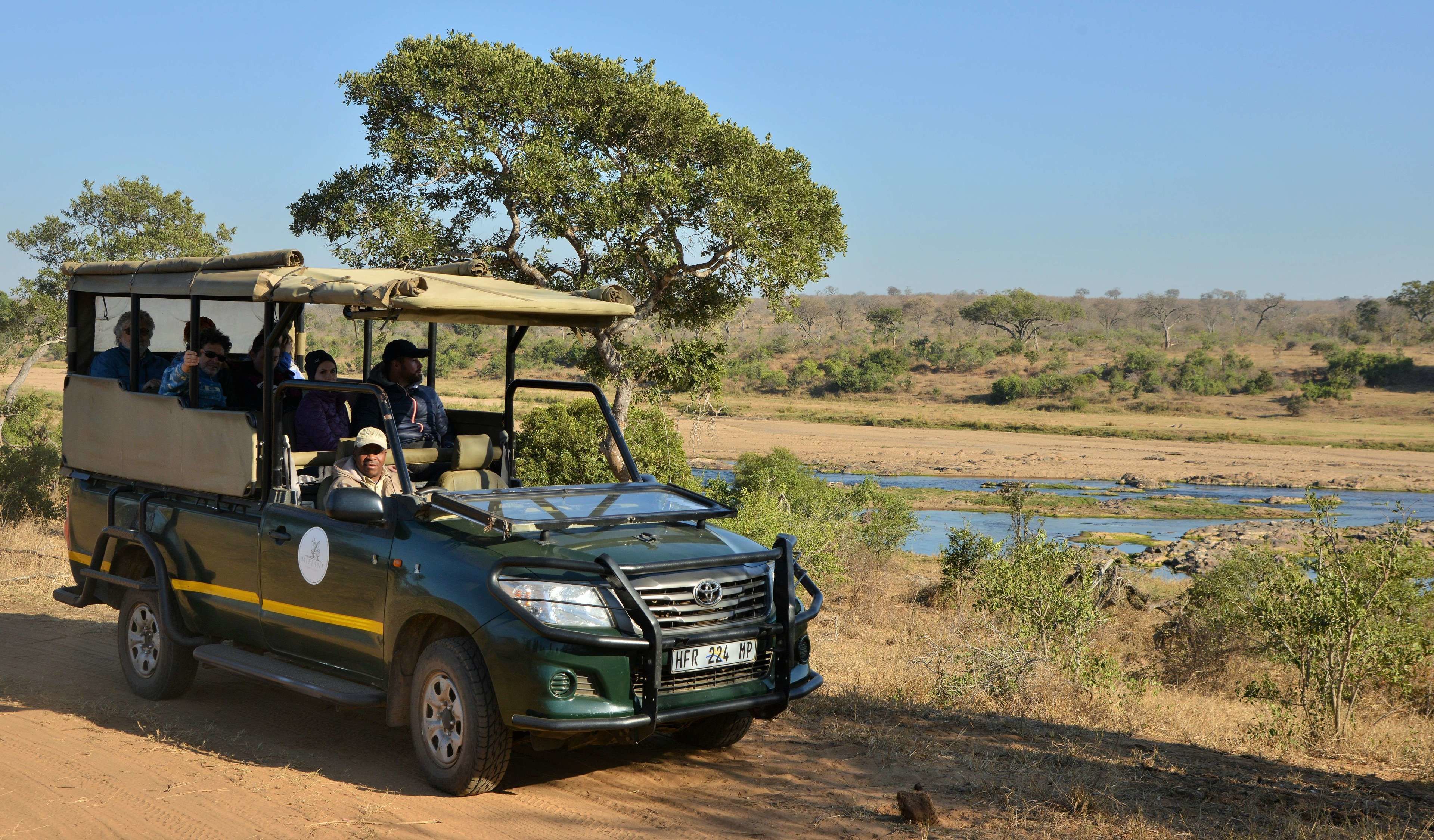 A safari vehicle with passengers, driving on a dirt road beside a river.