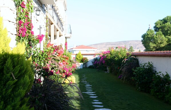 A garden pathway leading to a building with a red-tiled roof, surrounded by lush greenery and flowering plants.