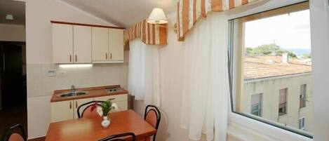 A compact kitchen with a dining table and chairs, a window with curtains, and a view of buildings outside.
