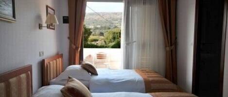 A hotel room with two beds, a window with curtains, a view of greenery outside, and a door.