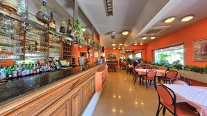 Angolo bar con scaffali pieni di bottiglie di liquori e bicchieri, un bancone e una zona con tavoli e sedie.