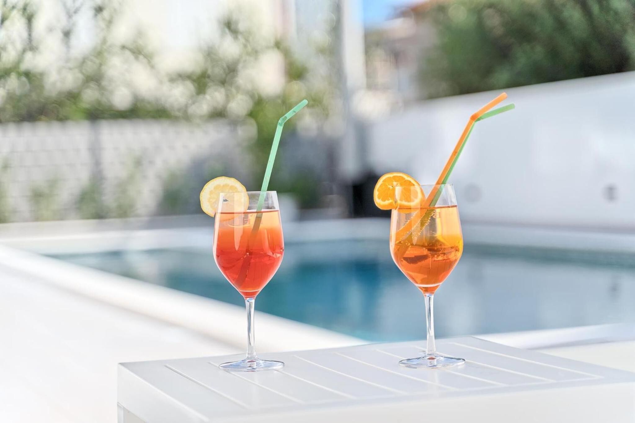 Two cocktails with straws and lemon slices on a table by a pool.