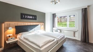A modern bedroom with a large bed, a nightstand with a radio, a wall-mounted light, and a window with a view of a garden. - Haus Granit Zermatt (Zermatt)