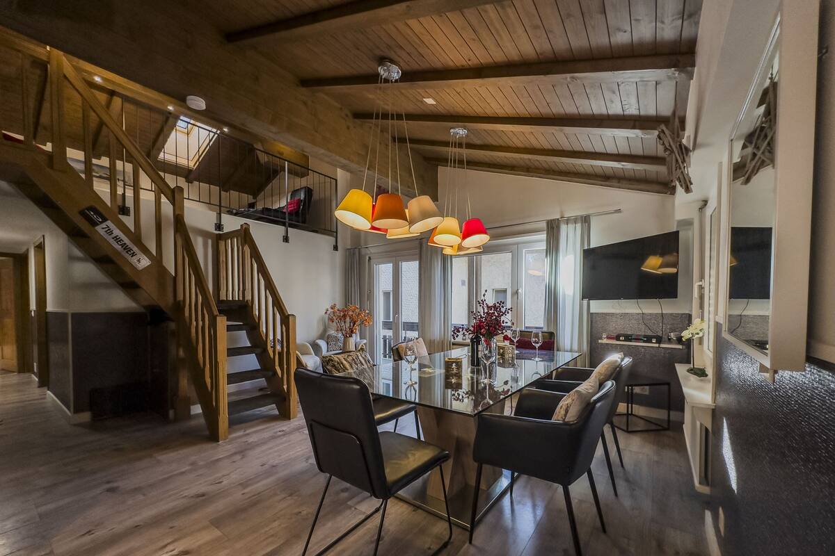 A spacious dining area with a wooden staircase, modern pendant lighting, and a view to the upper floor.