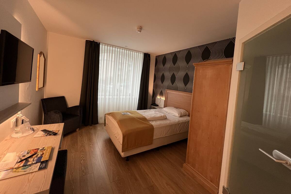 A hotel room with a bed, a desk with a chair, a TV mounted on the wall, and a window with curtains.