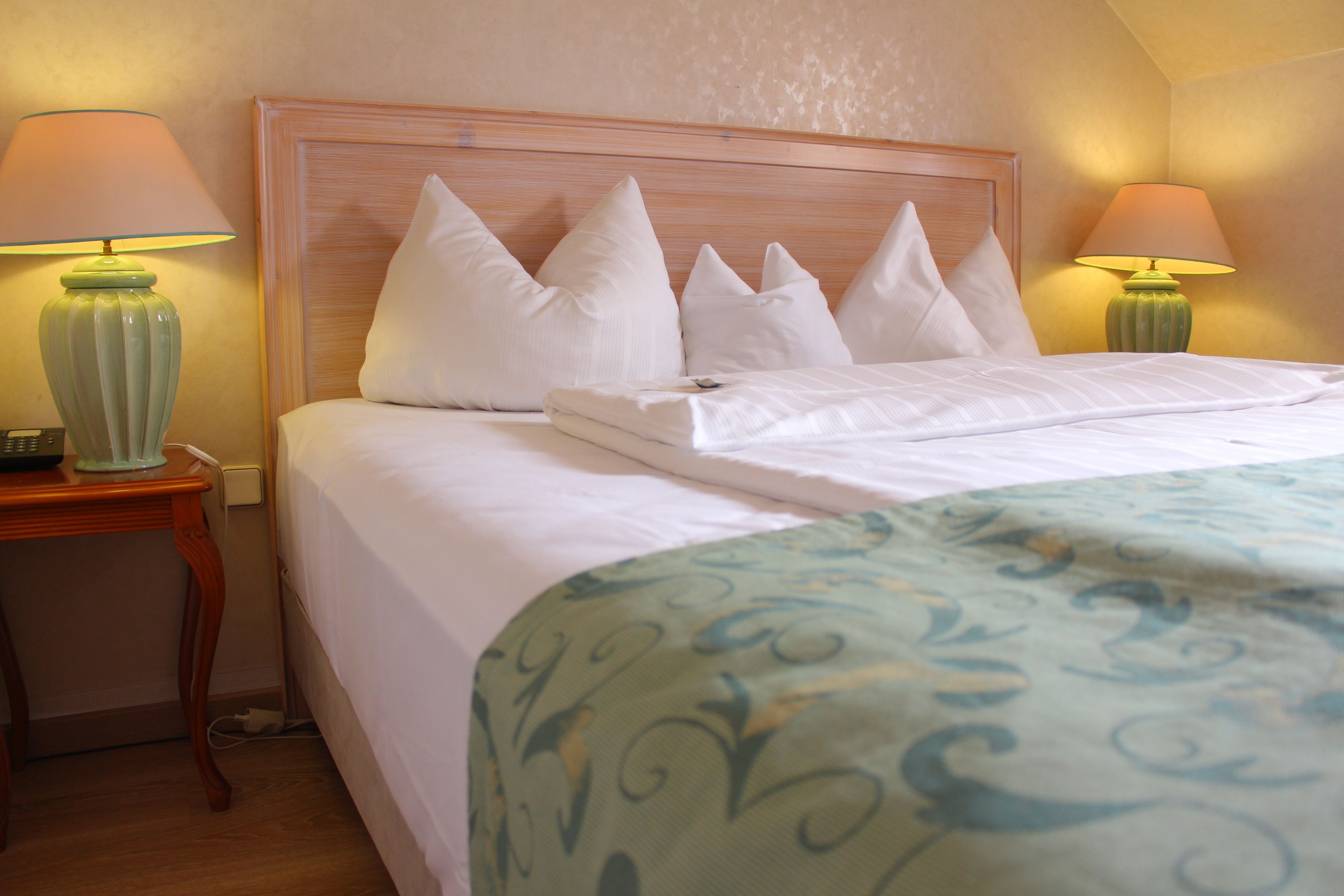 A neatly made bed with white linens and a blue patterned throw, flanked by two green lamps on wooden nightstands.