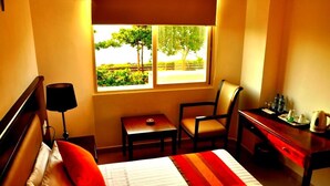 A hotel room with a bed, a desk, a chair, a window with a view of greenery, and a lamp. - Sai Sea City Hotel (Colombo)
