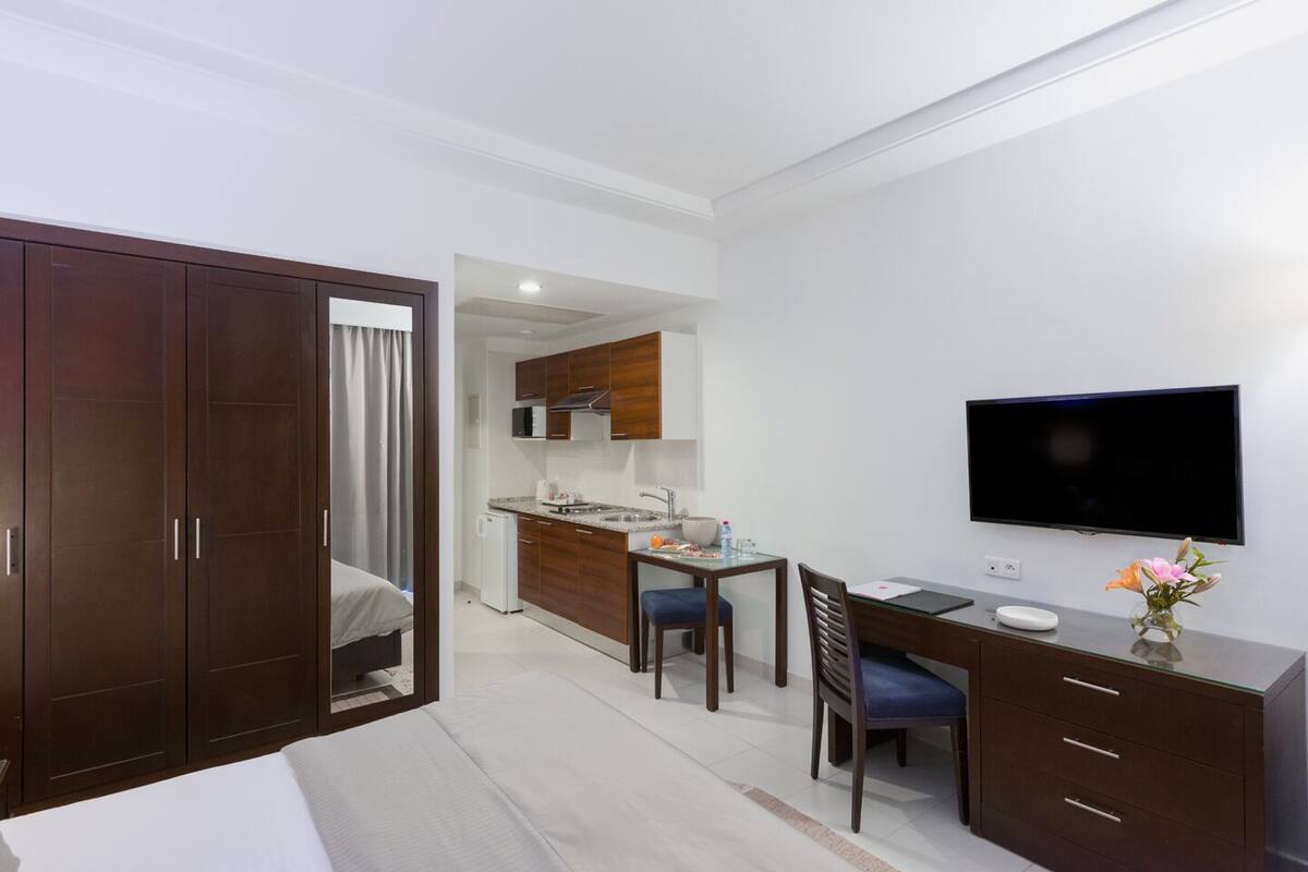 A hotel room with a bed, a desk with a chair, a TV, and a kitchenette.