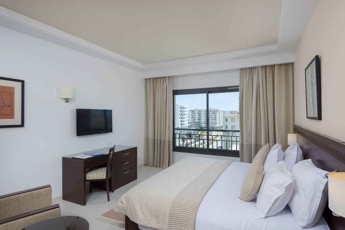 A hotel room with a bed, a desk, a chair, a television, and a window with a view of buildings.