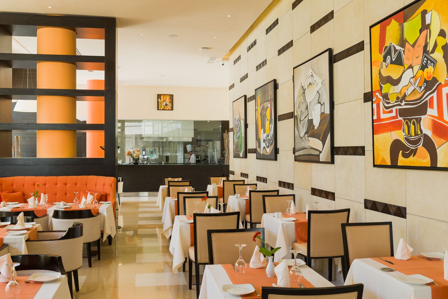 Een restaurant met meerdere tafels en stoelen, een centraal oranje zitgedeelte en kunstwerken aan de muren.