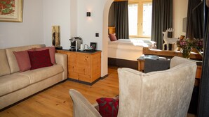 A cozy living room with a sofa, a wooden coffee table, and a chair. There is a bed with a window and curtains in the background. - Hotel Parsenn (Davos)