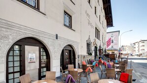 An outdoor seating area with wicker chairs and tables, a building with a sign reading "Hotel Barfenn", and a street view with other buildings and a sign for a restaurant. - Hotel Parsenn (Davos)
