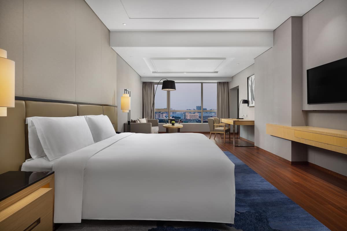 A modern hotel room with a large bed, a desk, a chair, a TV, and a view of the city.