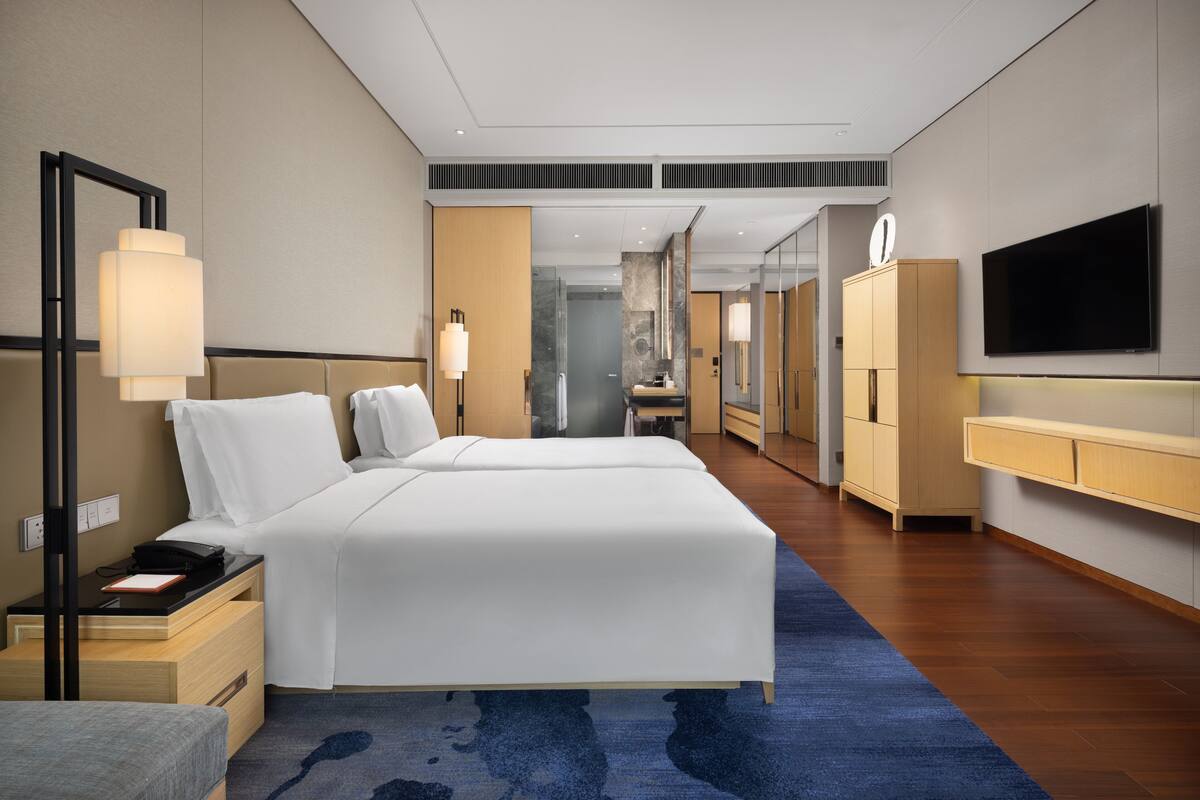 A modern hotel room with a large bed, bedside tables, a desk, a mirror, a TV, and a bathroom visible through a glass door.