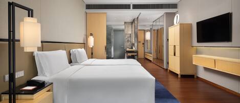 A modern hotel room with a large bed, bedside tables, a desk, a mirror, a TV, and a bathroom visible through a glass door.