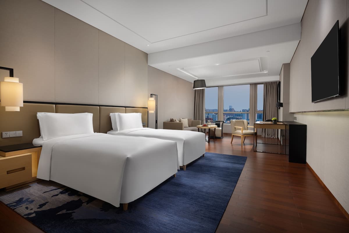 A hotel room with two beds, a desk, a chair, and a large window offering a city view.