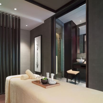 A modern spa room with a massage table, a small seating area, a wall-mounted mirror, and a large window.