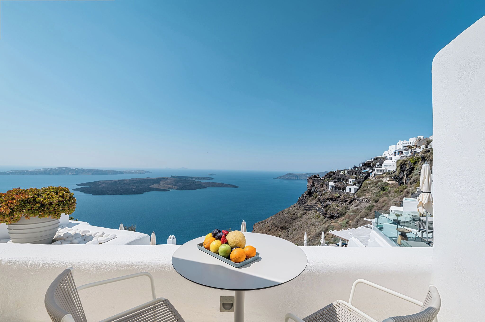 Un balcon avec une table dressée, ornée de fruits, d’où l’on peut admirer une vue sur la côte, avec ses bâtiments blancs et la mer.