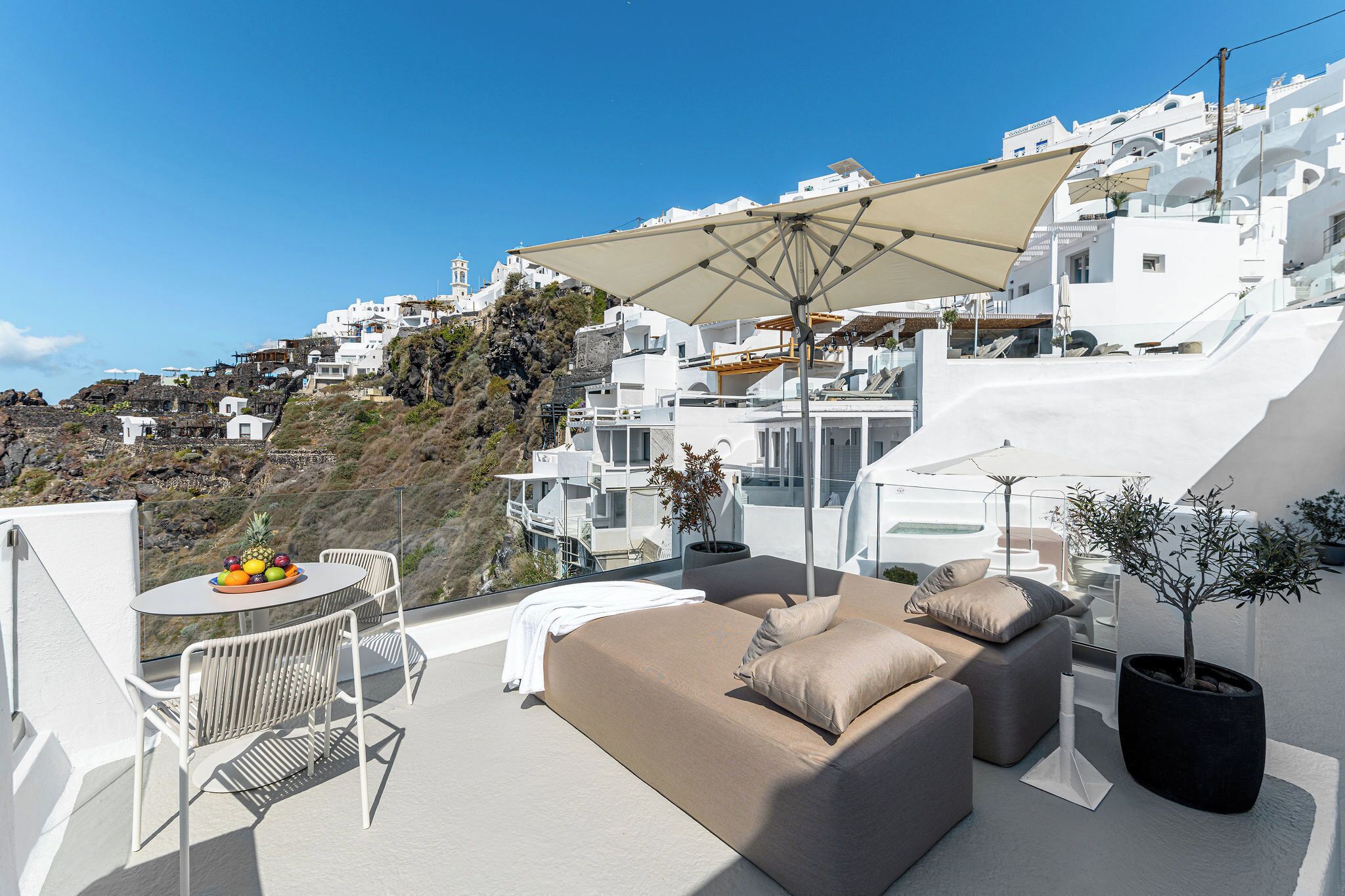 A modern outdoor balcony with a sofa, dining table, and umbrella, overlooking a white-washed hillside town.