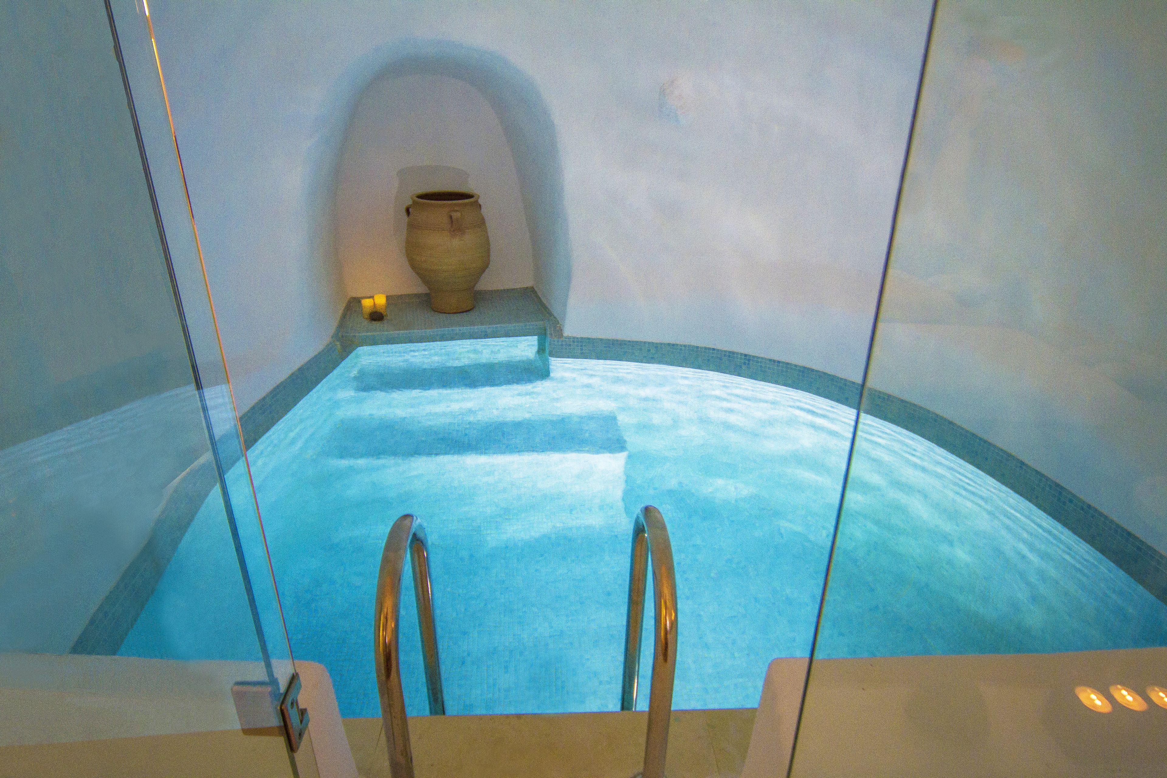 A clear blue pool with a ladder leading into it, a stone vase on a ledge, and a curved wall.